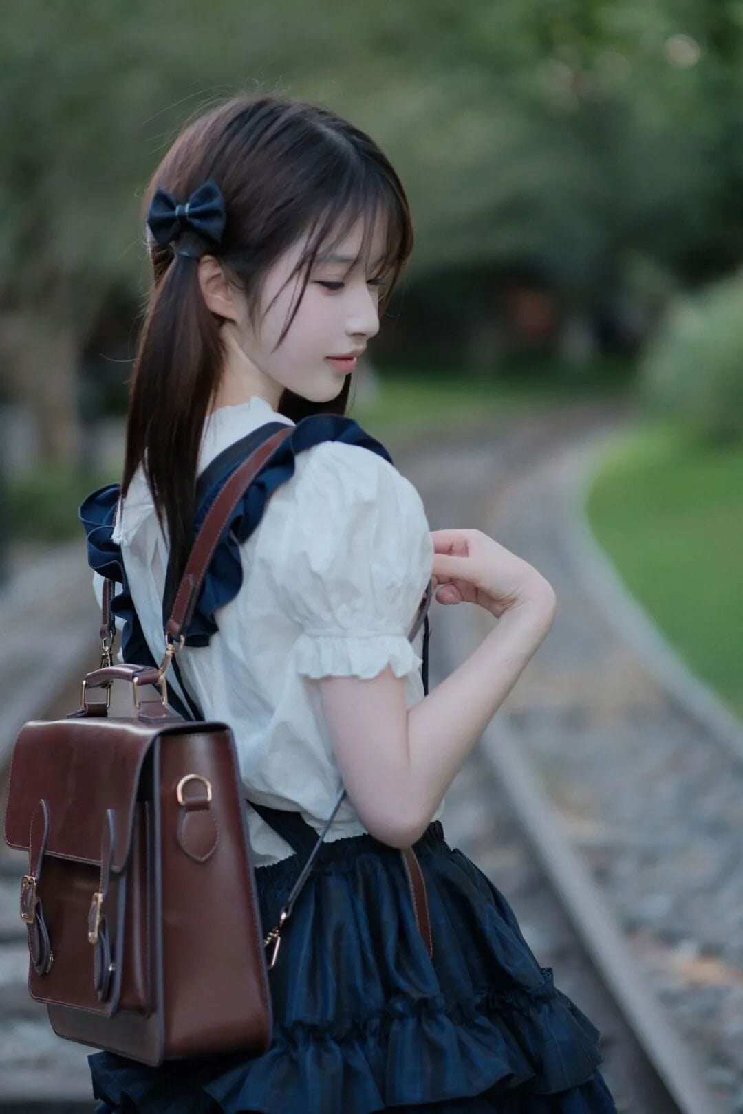 Close-up back profile of a girl wearing a dark brown vintage leather satchel backpack near train tracks