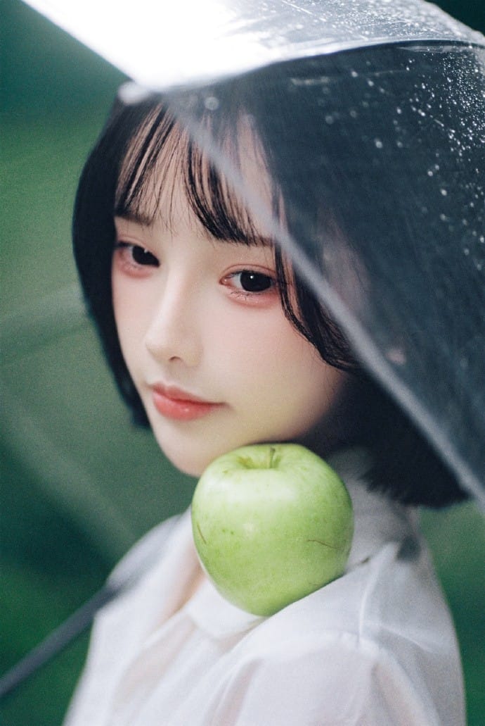 Cinematic close-up portrait of Ah Xuan resting a green apple on her shoulder under a raindrops covered umbrella