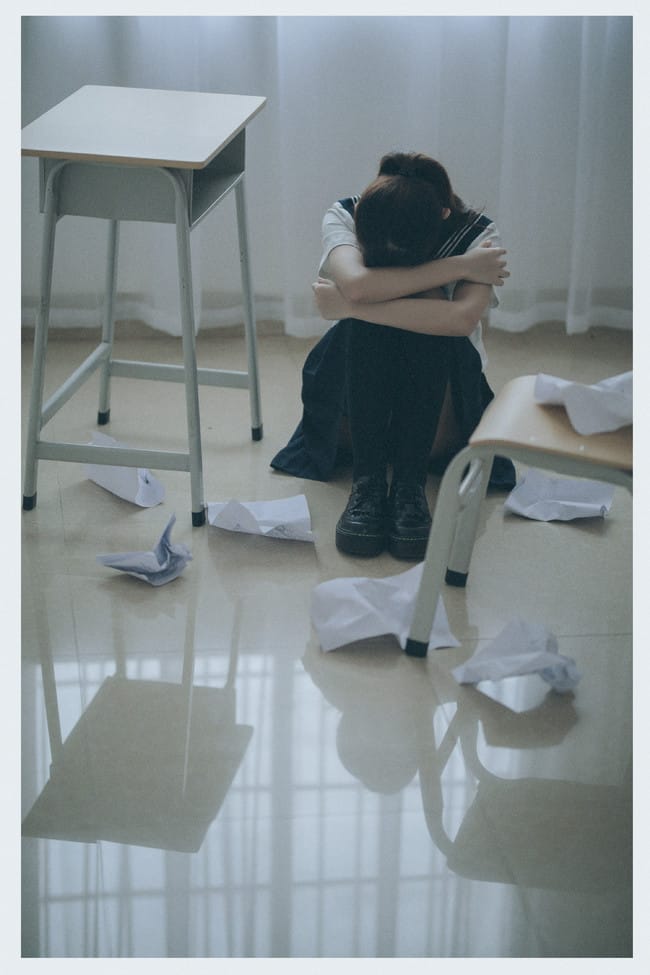 Sukii阿蛊 curling up on the floor surrounded by crumpled papers and empty classroom chairs