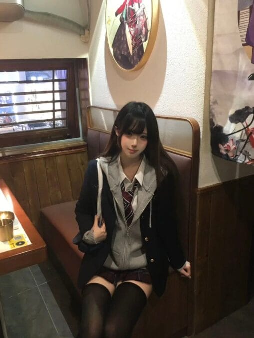 Girl in navy blazer, grey zip hoodie, red necktie, and plaid skirt sitting in an izakaya wooden booth
