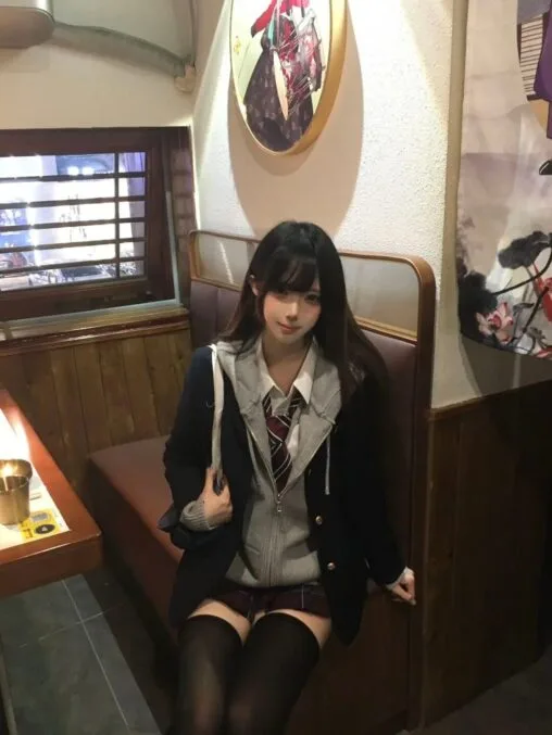 Girl in navy blazer, grey zip hoodie, red necktie, and plaid skirt sitting in an izakaya wooden booth